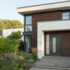 Moderne Haustür mit großen Fenstern und Holzverkleidung Ein modernes Haus mit einer dunklen Haustür, großen Fenstern und einer Holzverkleidung an der Fassade. Der Eingang ist von einem kleinen Garten umgeben.